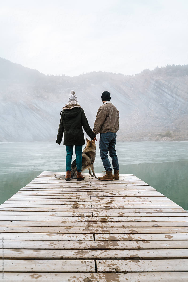 Couple on wooden dock in frozen lake in mountain with dog
