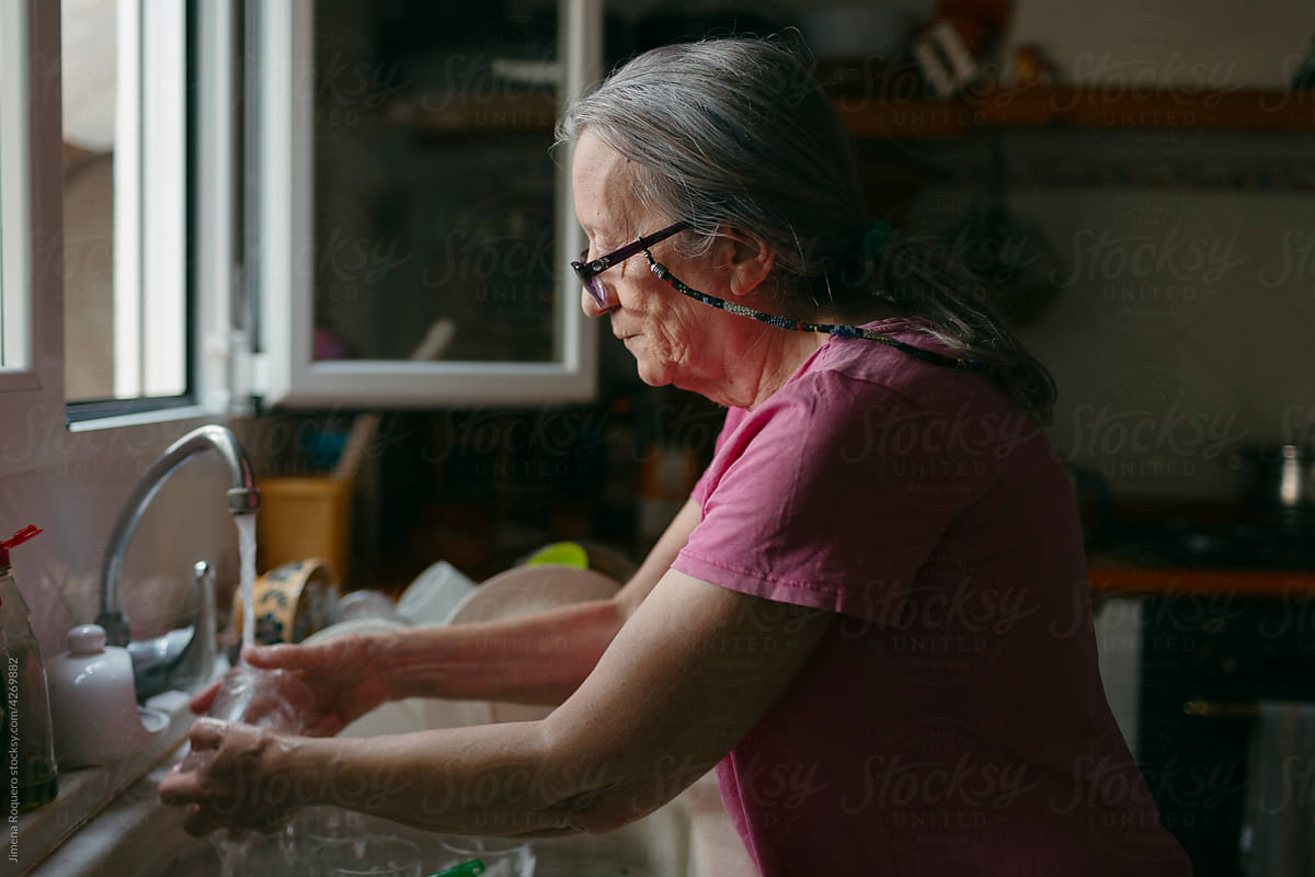 Elderly grey-haired woman doing dishes by a window