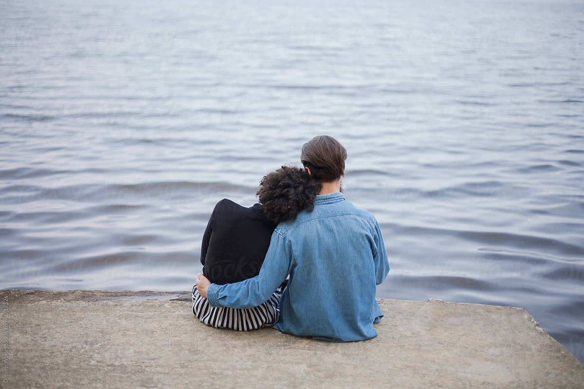 photo of a hugging couple from behind. couple sitting on a pier against the background of water