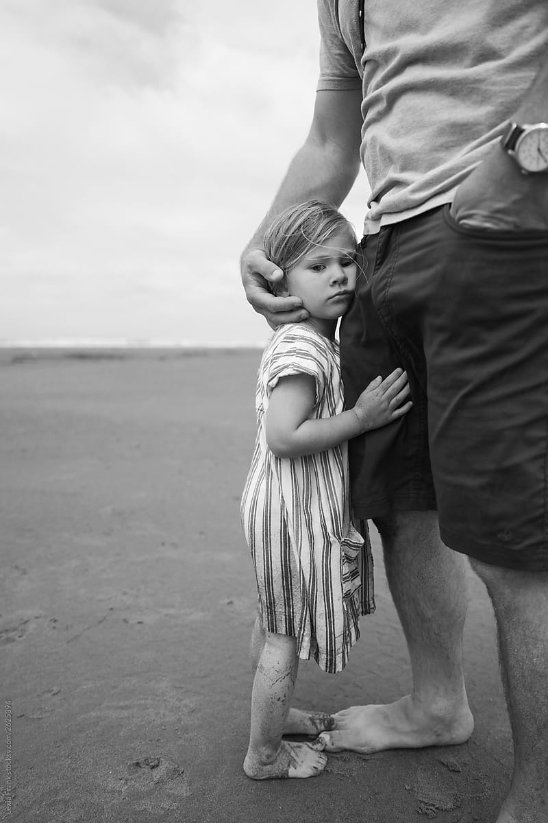 sad, tired little girl holds on to daddy\'s leg at the beach with dirty legs
