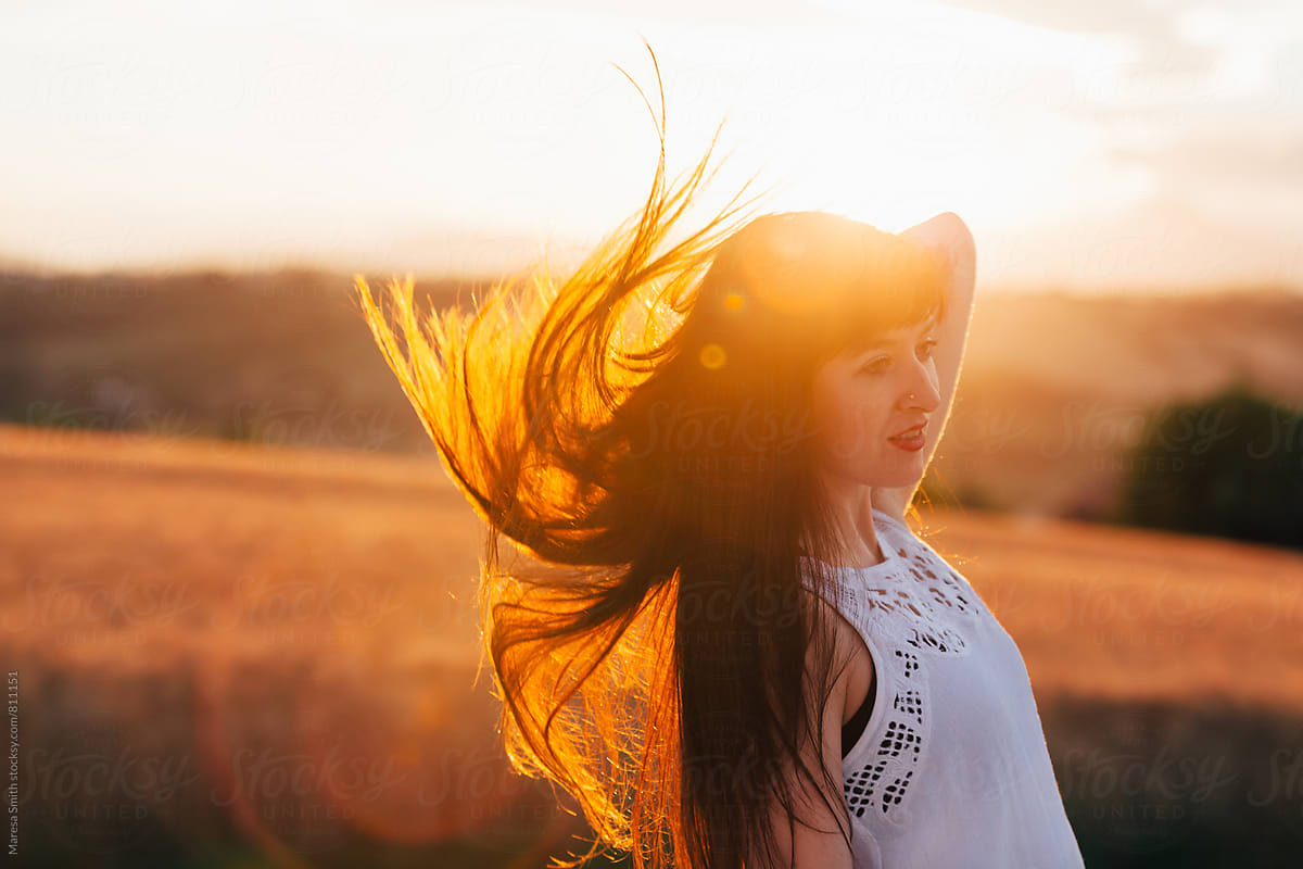 A brunette lady flicks her hair back, it is back lit by the sunset