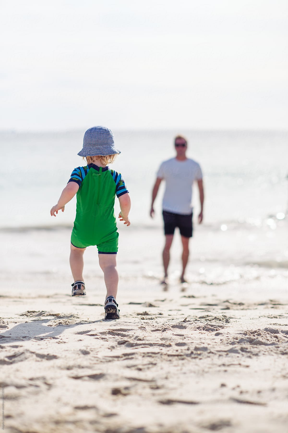Toddler in swimsuit and hat walks towards his father at beach