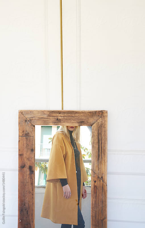 Girl in yellow coat reflecting in mirror over white wall.