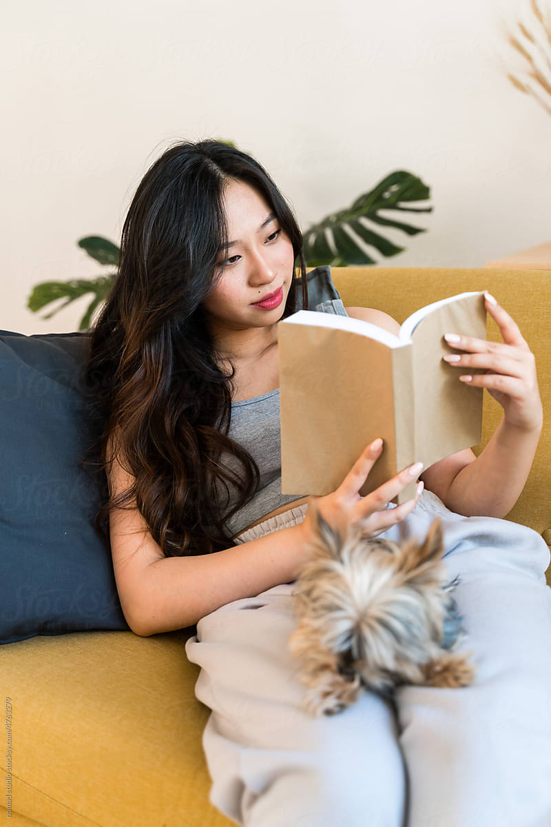 Woman reading a book sitting on sofa with her dog.