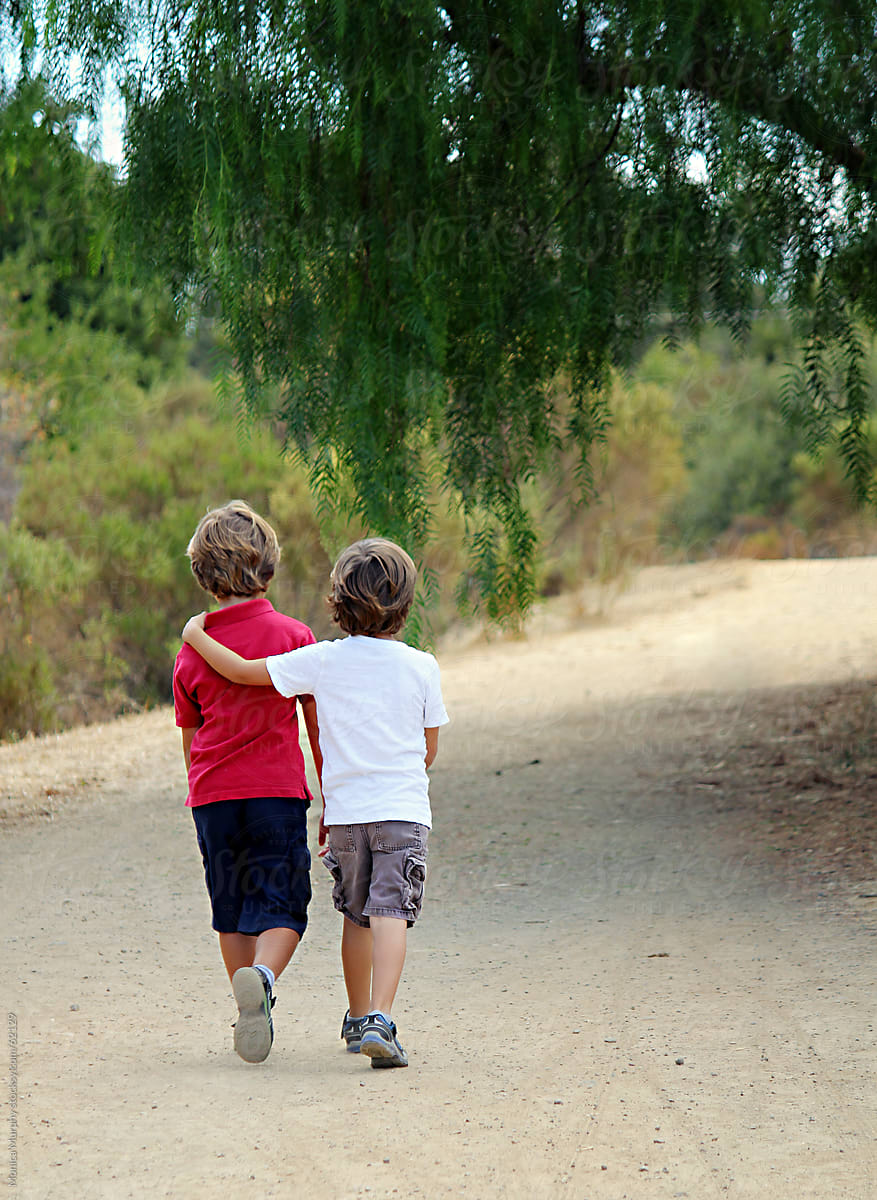 Two boys walking down trail, one with arm around the other, leading into  the trees by Monica Murphy - Stocksy United
