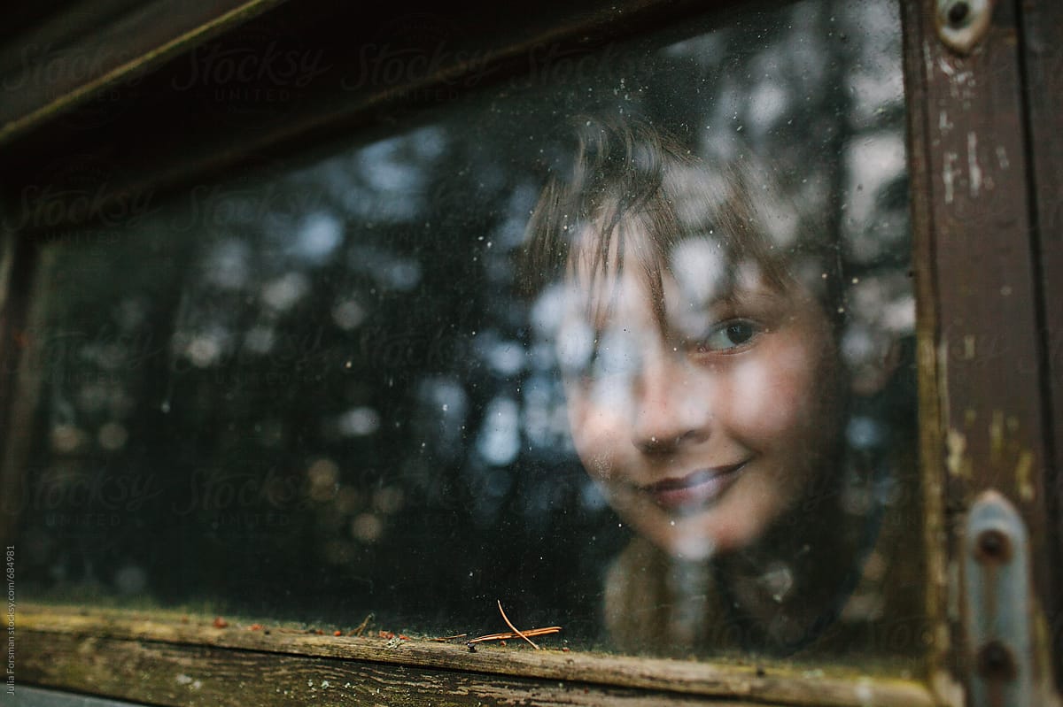 Boy looks through the weathered window of a small window in a wooden building.