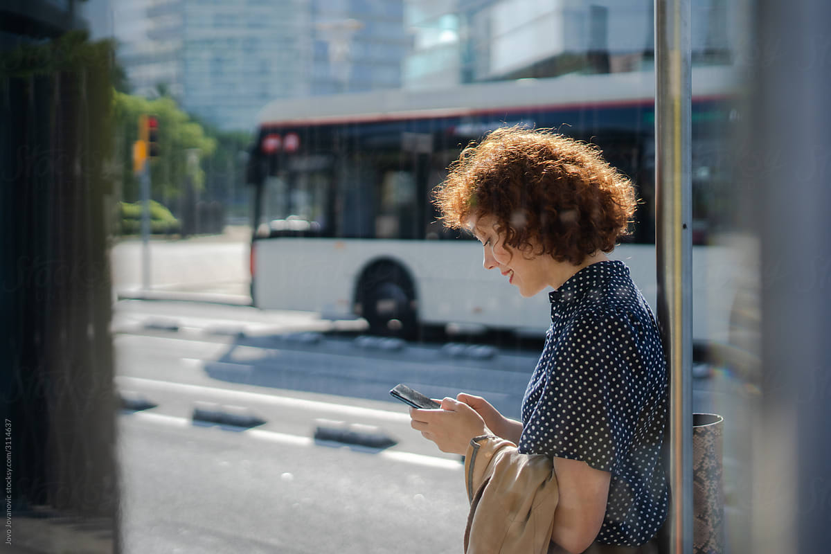 Curly redhead woman answering messages on phone while waiting for bus