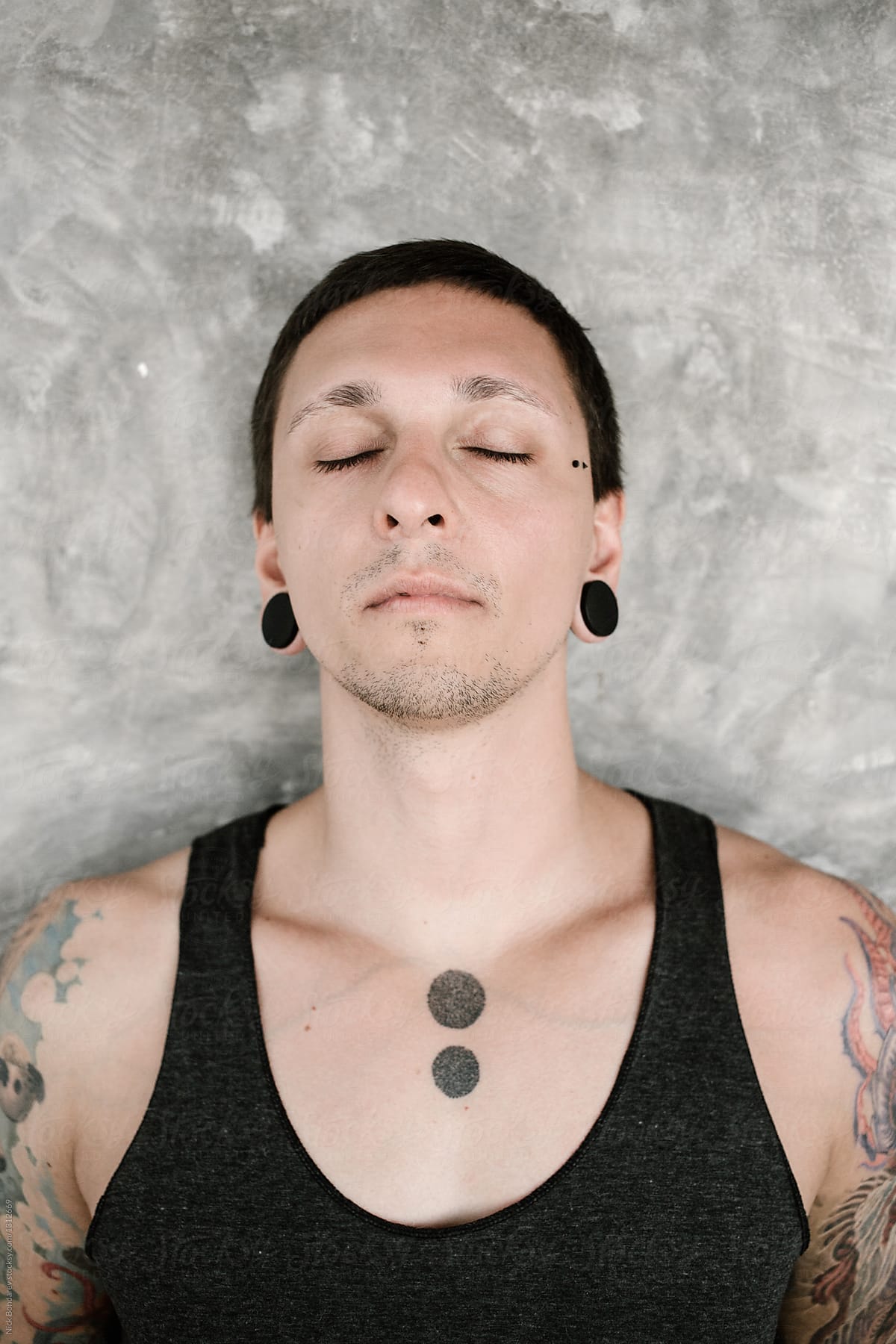 Portrait of young man with tunnels and colourful tatoos with closed eyes on gray wall background