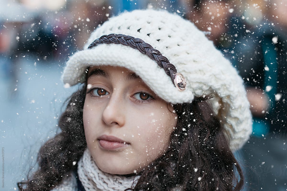Headshot of a girl looking at camera wearing a white cap in the winter with snowflakes
