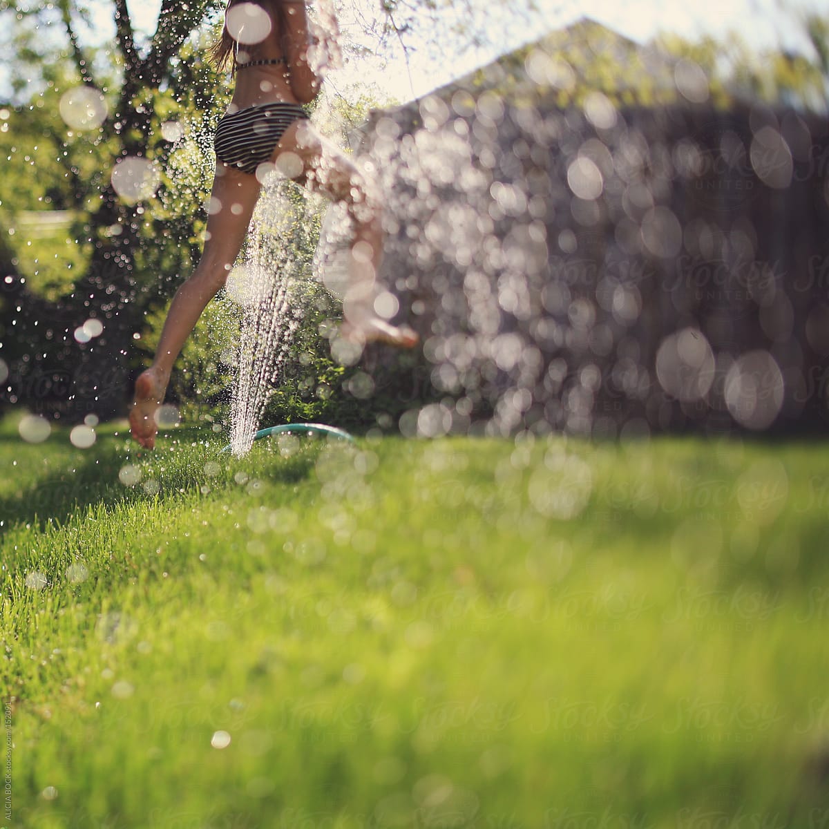 Girl Having Fun And Running Through The Sprinkler On A Hot Summer Day