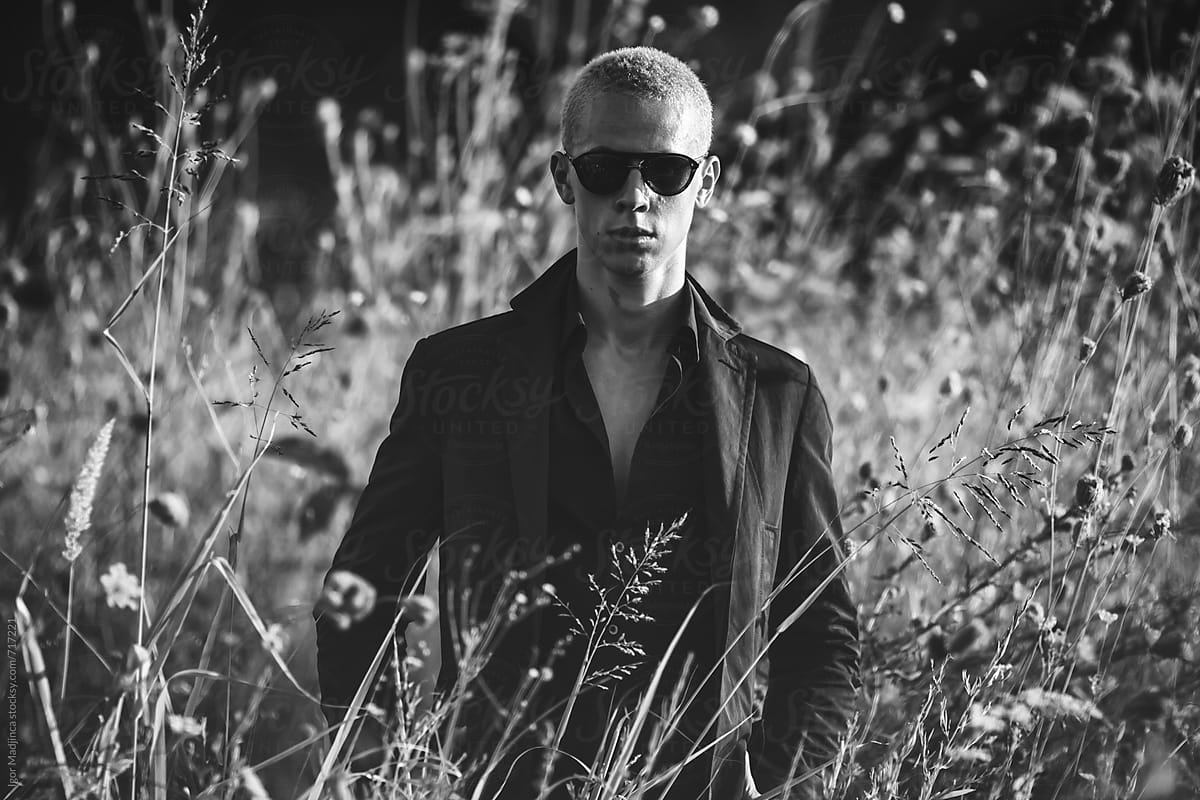 blond young man with black glasses in a field of tall grass, dressed in a black
