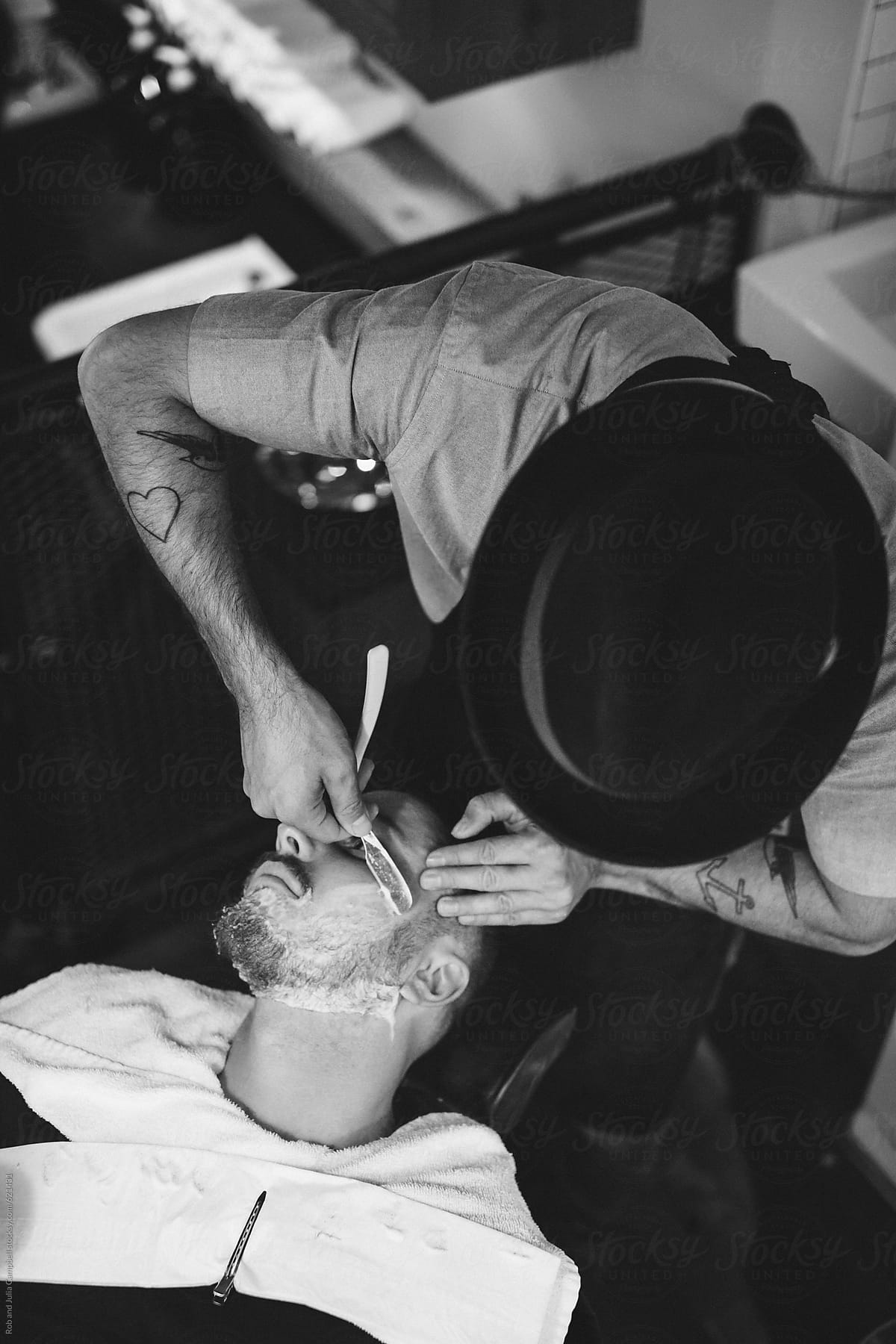 Hat-wearing barber giving man a classic hot lather shave - top view