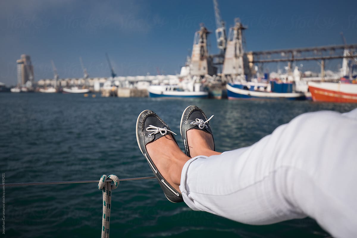 Young woman wearing boat shoes with laces tied and feet back relaxing on yacht floating on the ocean with boats in harbour port