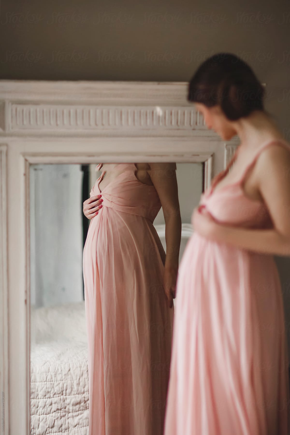 Woman touching and looking in mirror her pregnant belly while wearing a pink dress