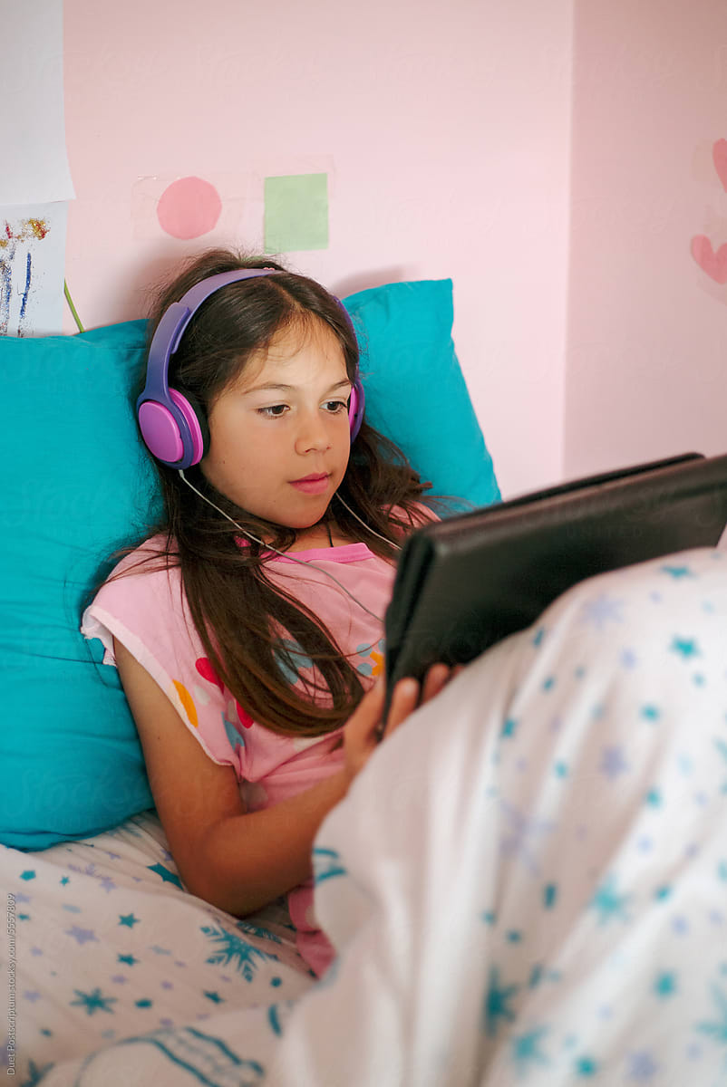 A little girl is looking at an electronic tablet lying in her bed