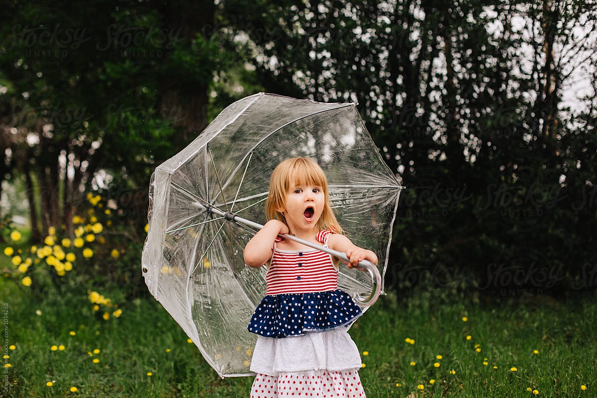 Toddler girl in an American flag dress holding a big, clear umbrella.