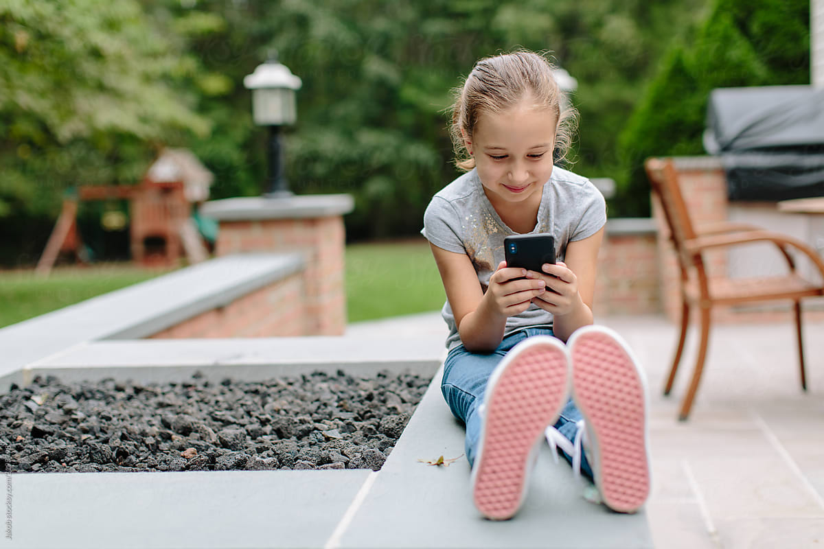 Cute Young Girl Holding A Phone And Smiling By An Outside Fireplace By Stocksy Contributor