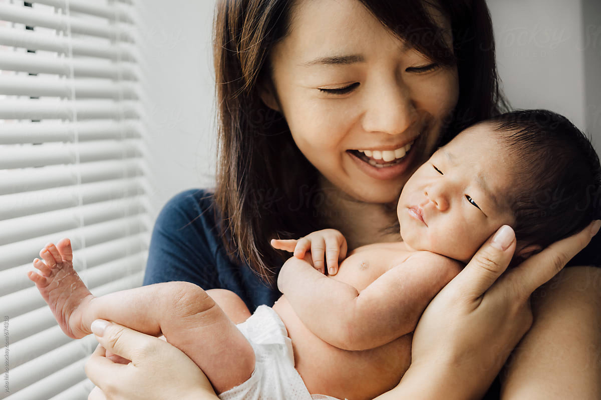 Happy Asian Mother Smiling At Her Newborn Baby Girl In Her Arms By
