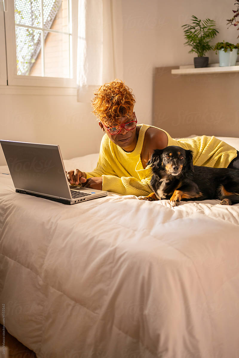 Black Woman With Laptop Stroking Her Dog On The Bed.