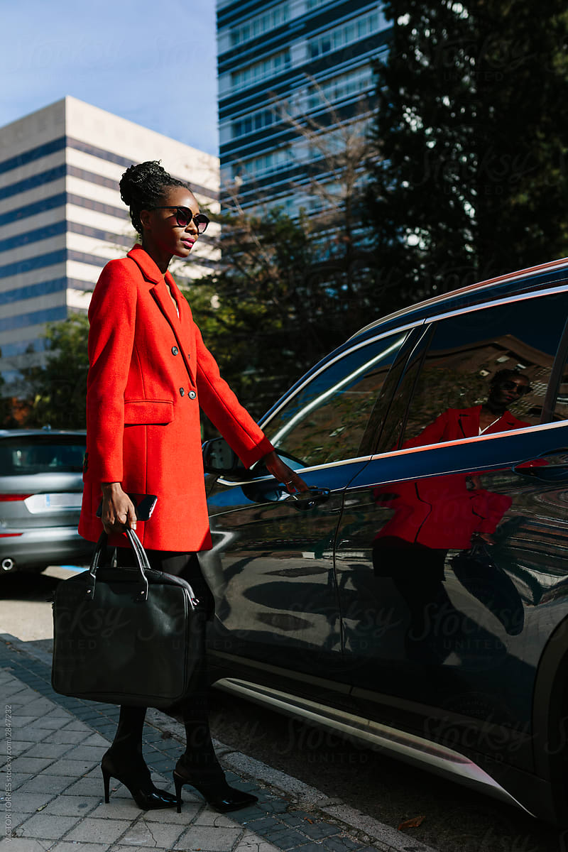 Active black woman in sunglasses and coat opening car door in street
