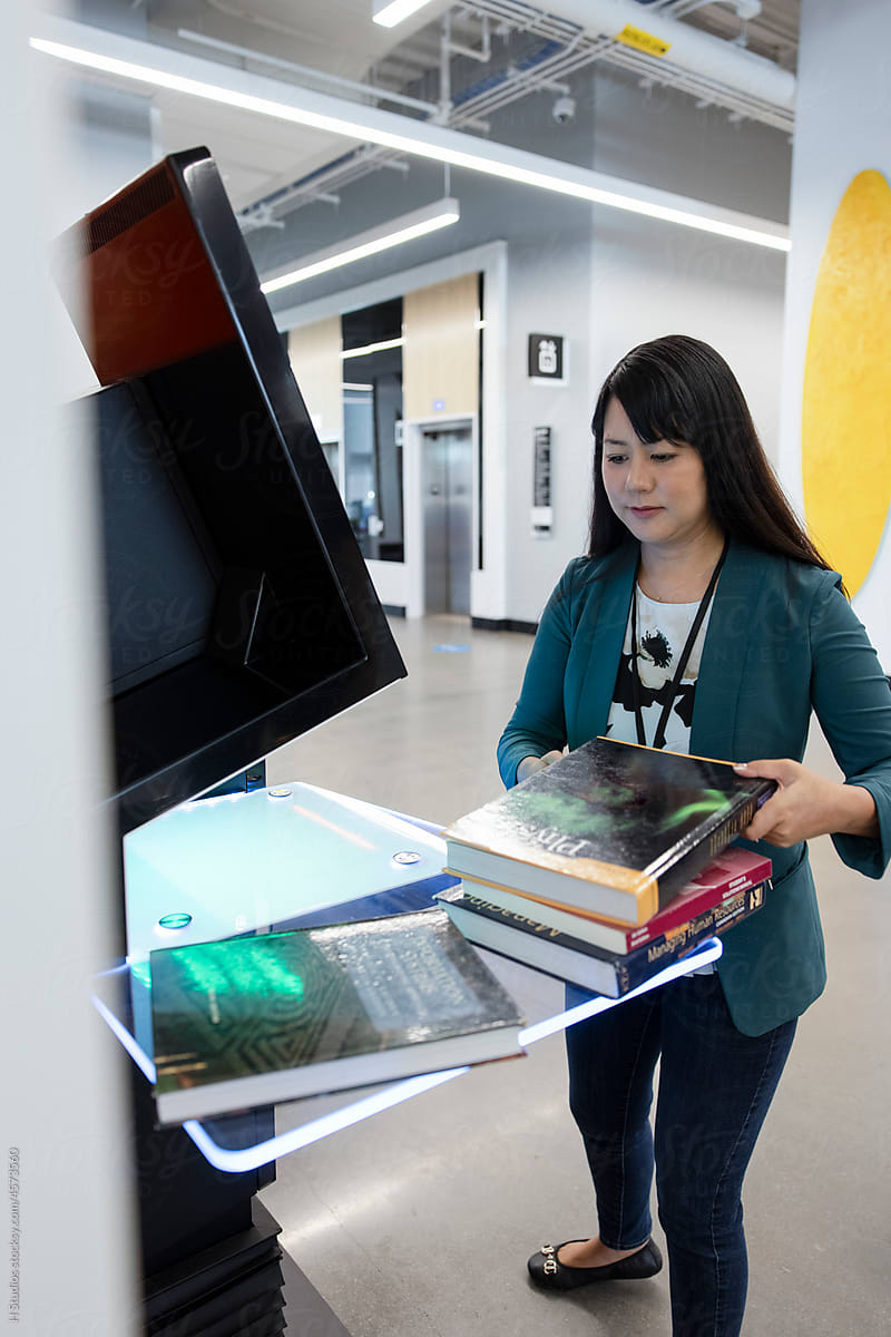 Female librarian with books using self checkout kiosk in library.