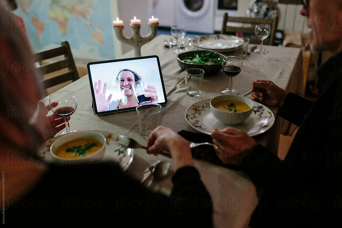 Woman virtually toasting on the screen of a device on the table while elderly couple have dinner.