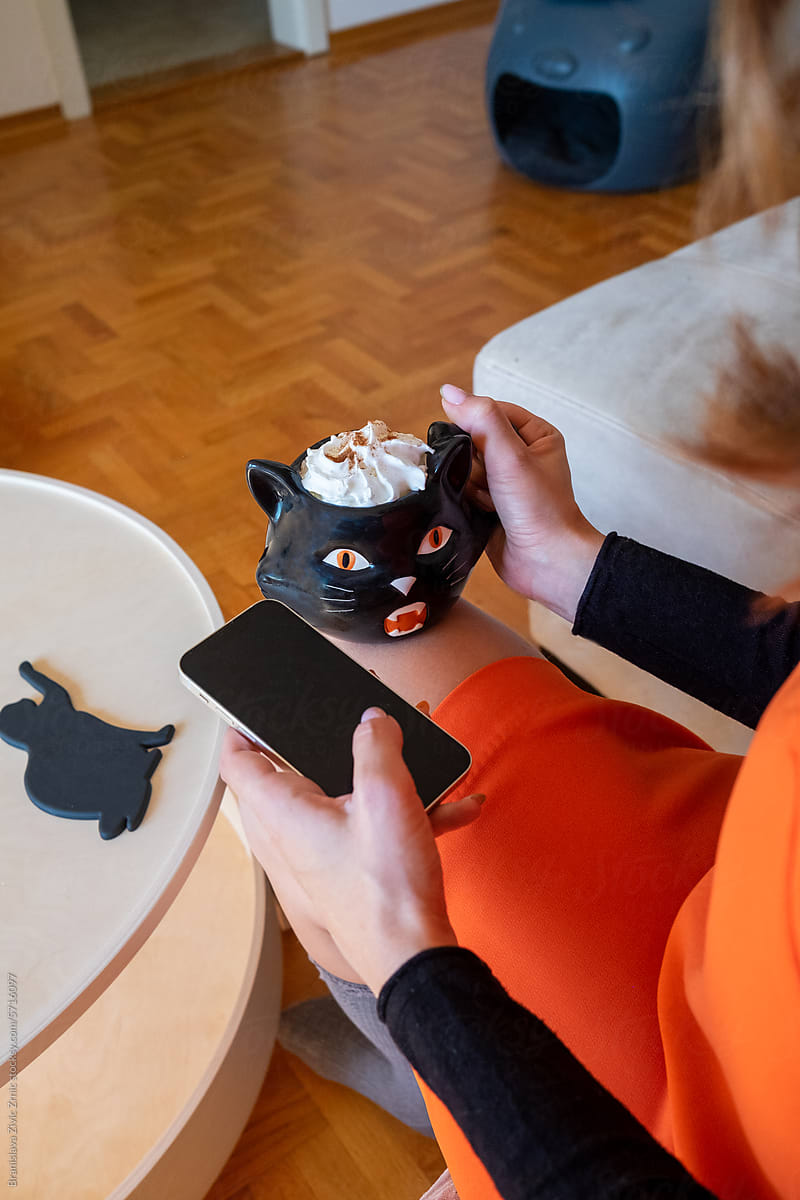 Female using her phone and drinking pumpking latte from halloween mug