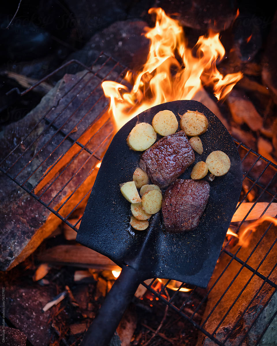 Steak and potatoes being cooked over an open fire in a shovel