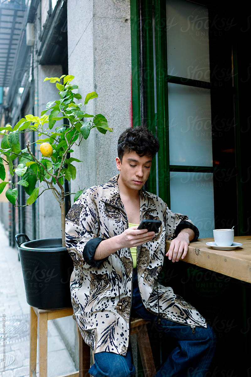Young extravagant gay man sitting outside of a city cafeteria checking his phone