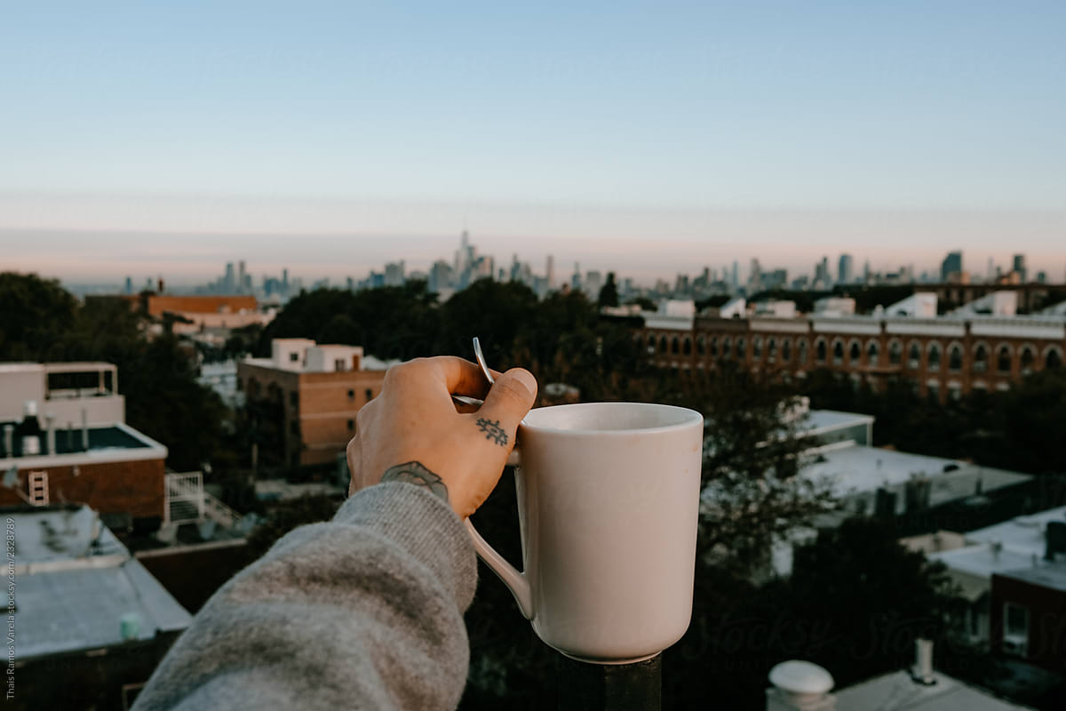 hand with a white cup of coffee looking towards the new york skyline