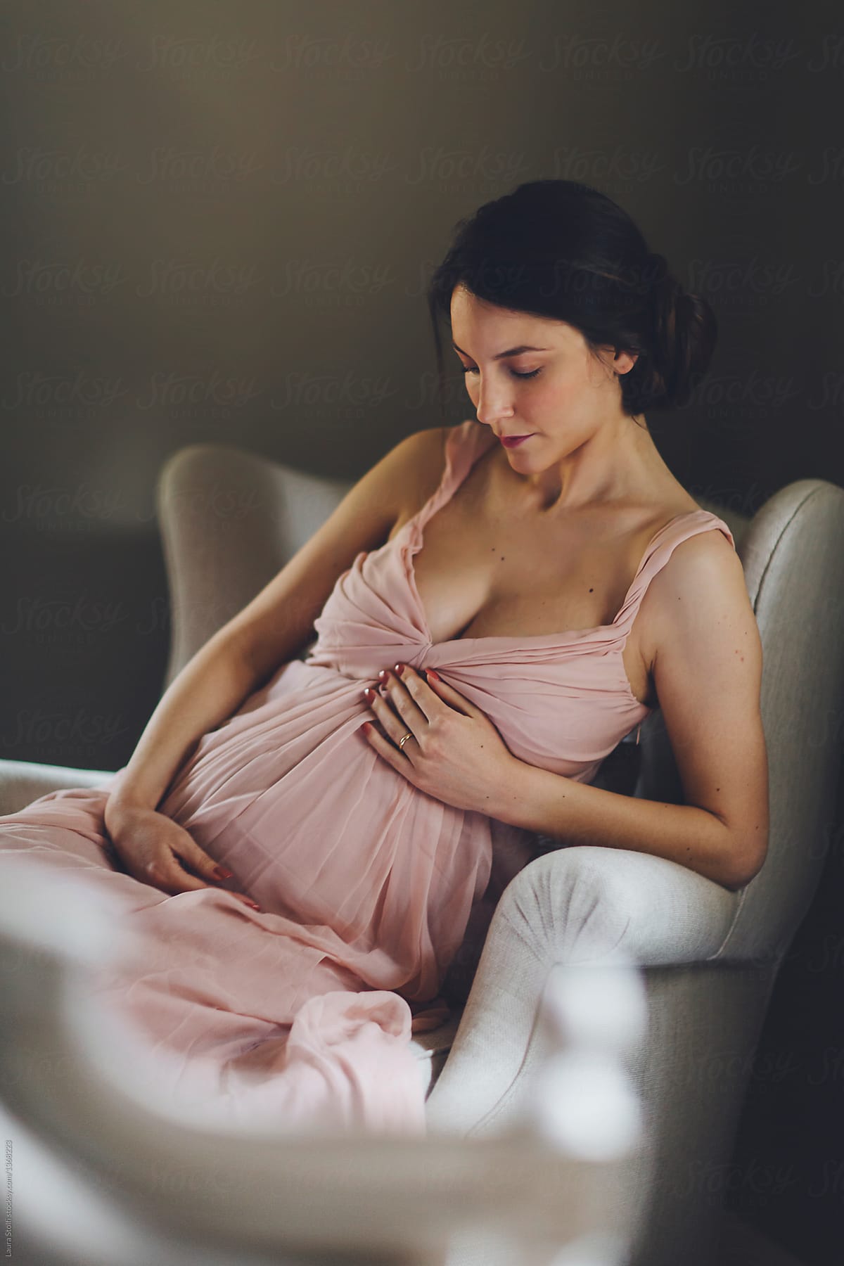 Pregnant woman sits on armchair wearing a beautiful pale pink dress by Laura Stolfi for Stocksy United