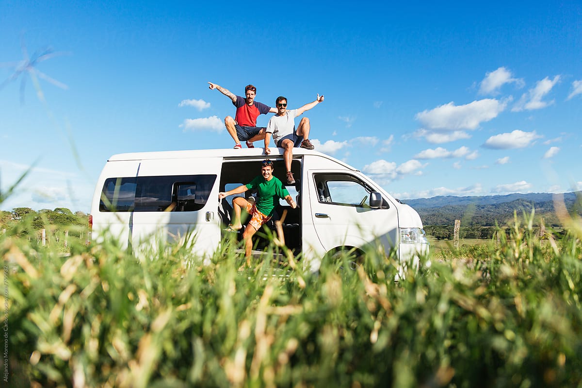 Three excited young men sitting on the roof of a white minivan during a break stopover in the middle of the road in the countryside with grass in front