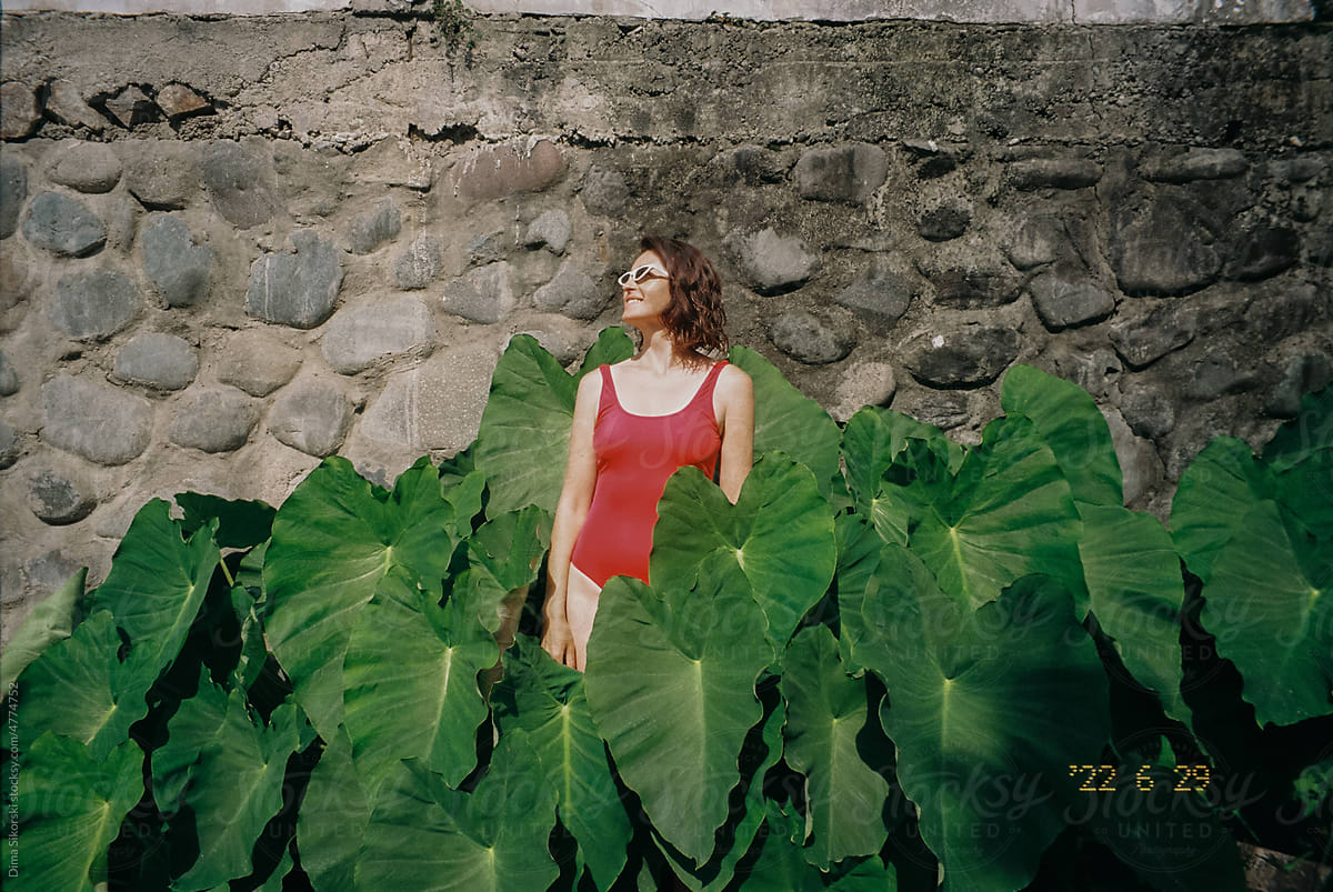 A curly-haired girl in a bright swimsuit smiles near huge green leaves