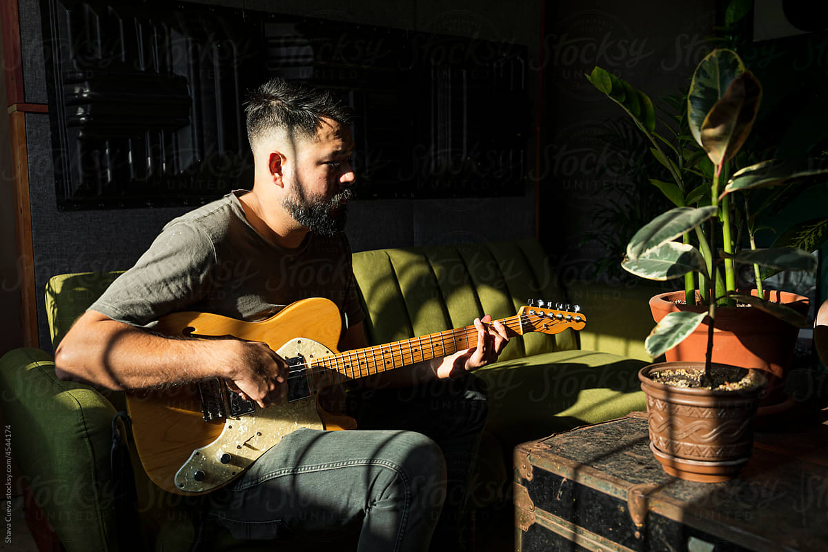 Man with beard playing an electric guitar in a room with plants