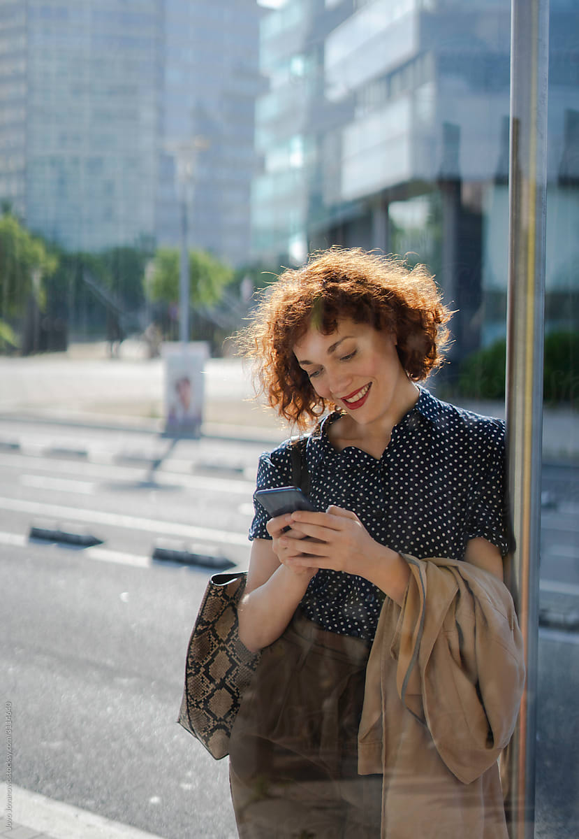 Smiling red headed woman waiting for bus with smartphone