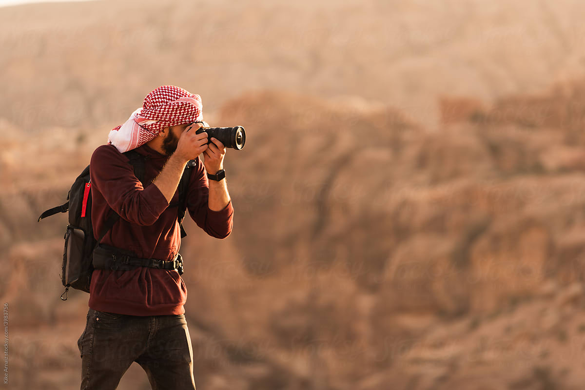 Man in keffiyeh shooting on camera valley of desert on top of cliff