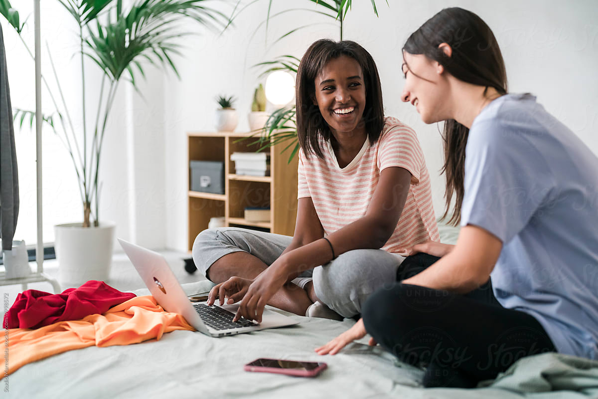 Smiling diverse female friends sharing laptop on bed