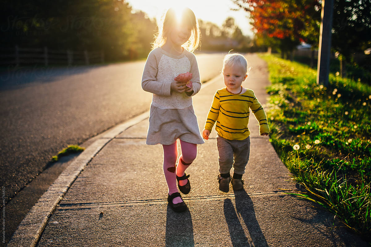 Young Girl and Her Toddler Brother Walk on the Sidewalk in the evening light