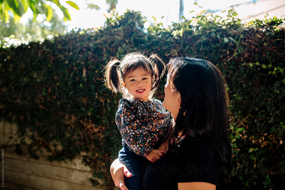 Smiling asian toddler girl in her mother\'s arms outside in backyard