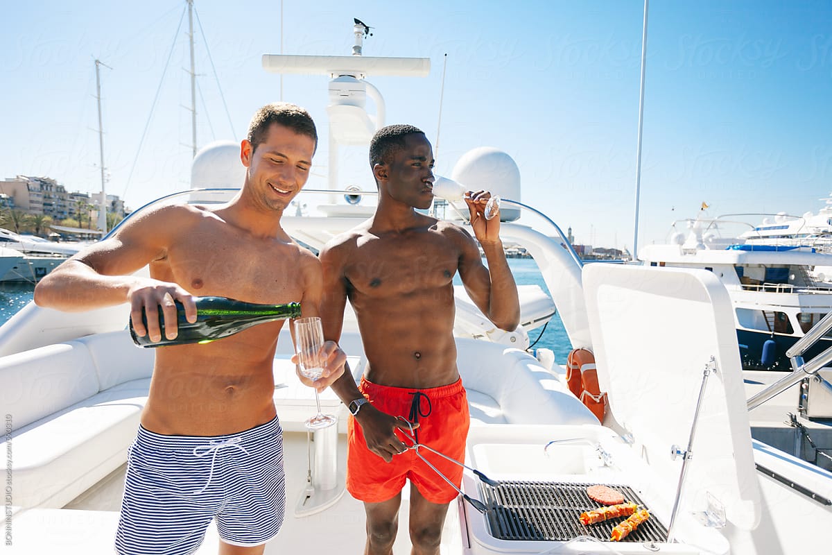 Young male friends drinking champagne whilst cooking meat in a grill on a luxurious yacht.