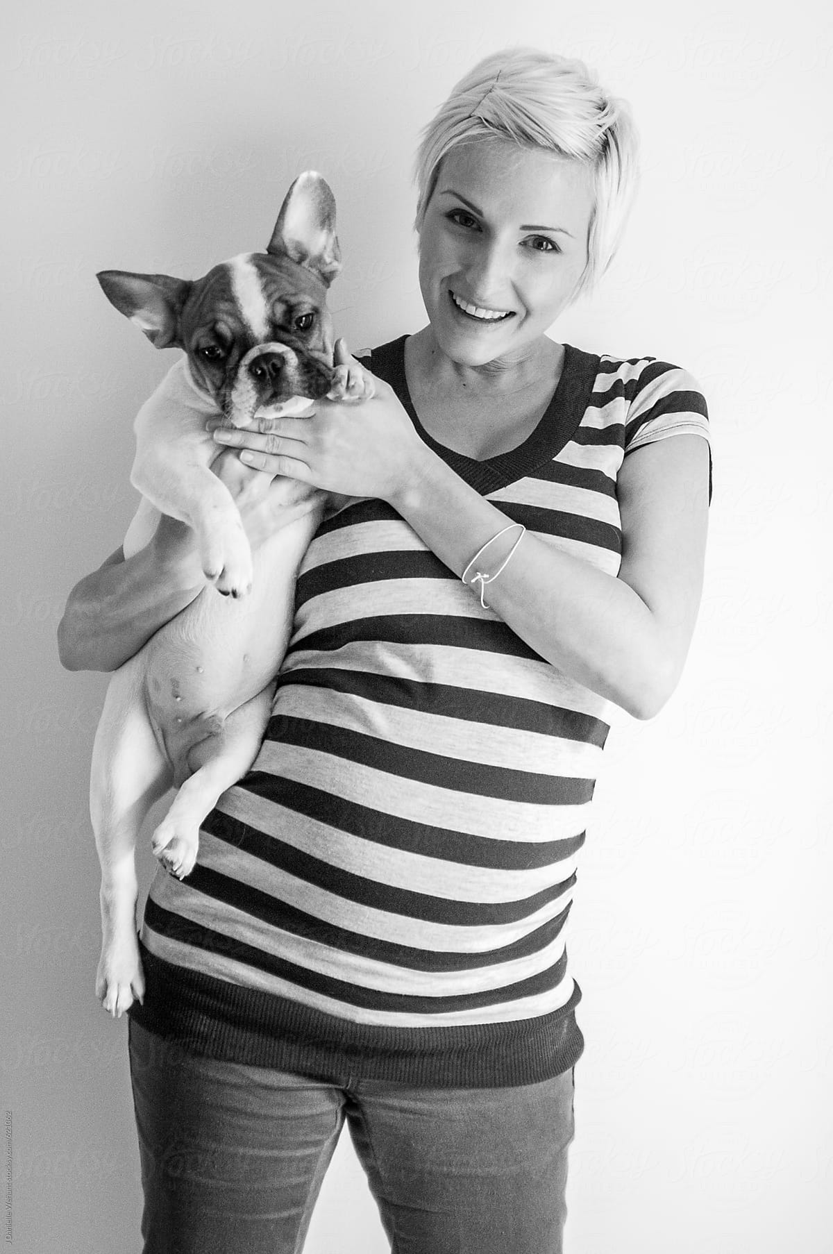 A pregnant caucasian woman with blonde hair holding a french bulldog puppy.