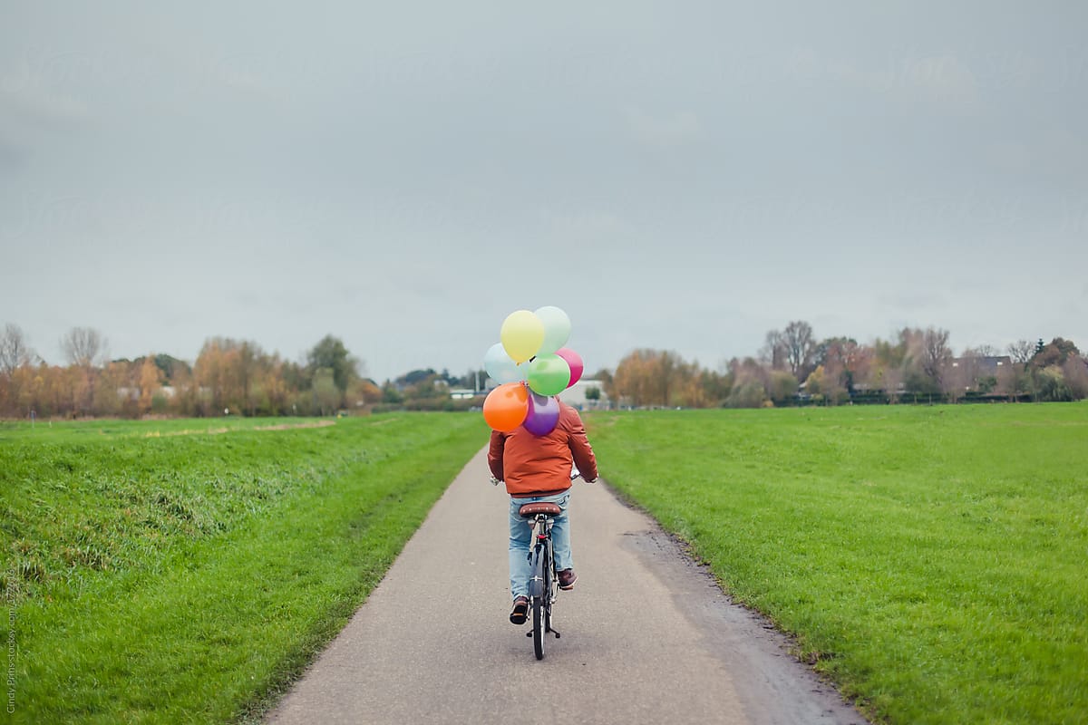 Teenage boy riding away on a bike holding a bunch of balloons