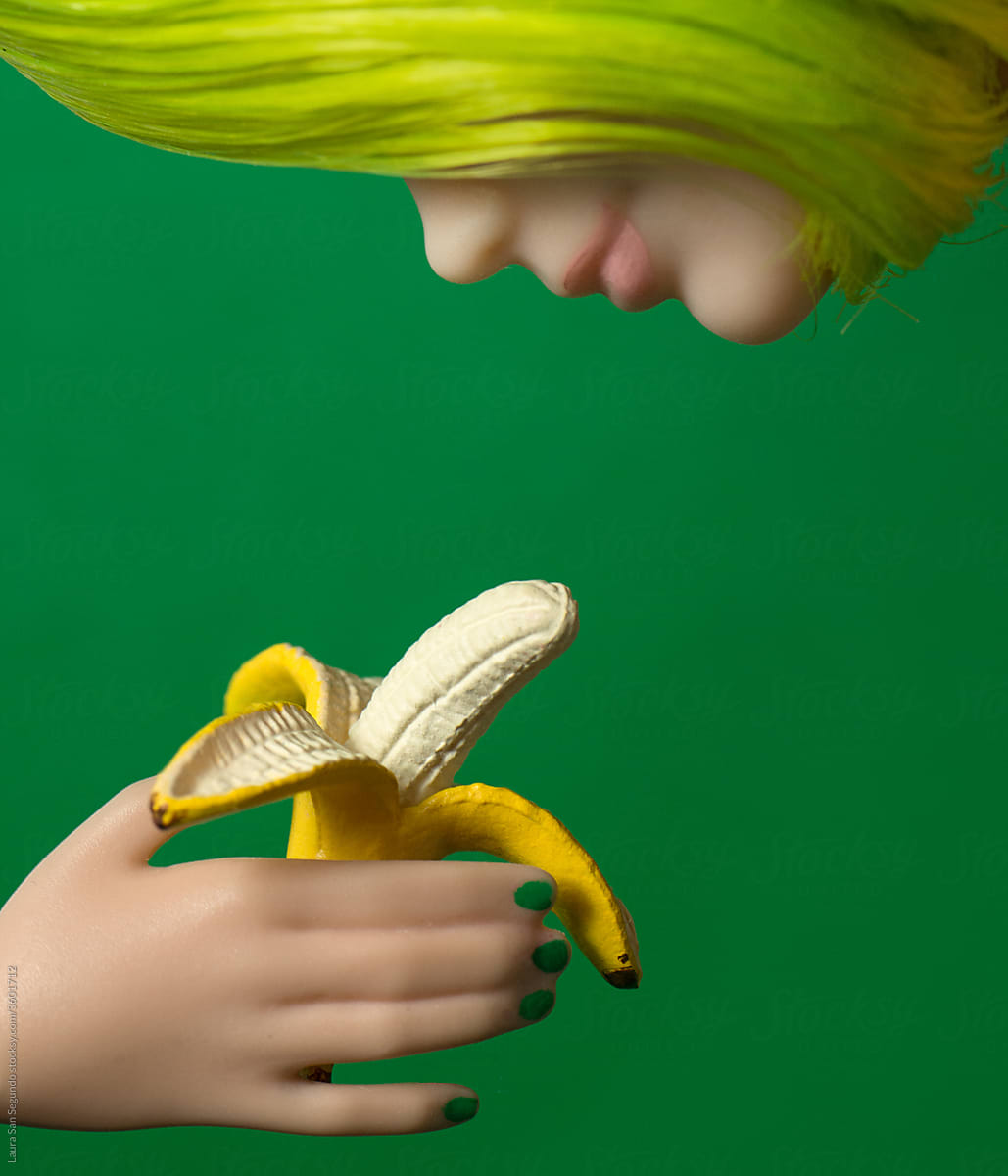 Plastic hand with green nails holding an open banana in front of a green haired doll.