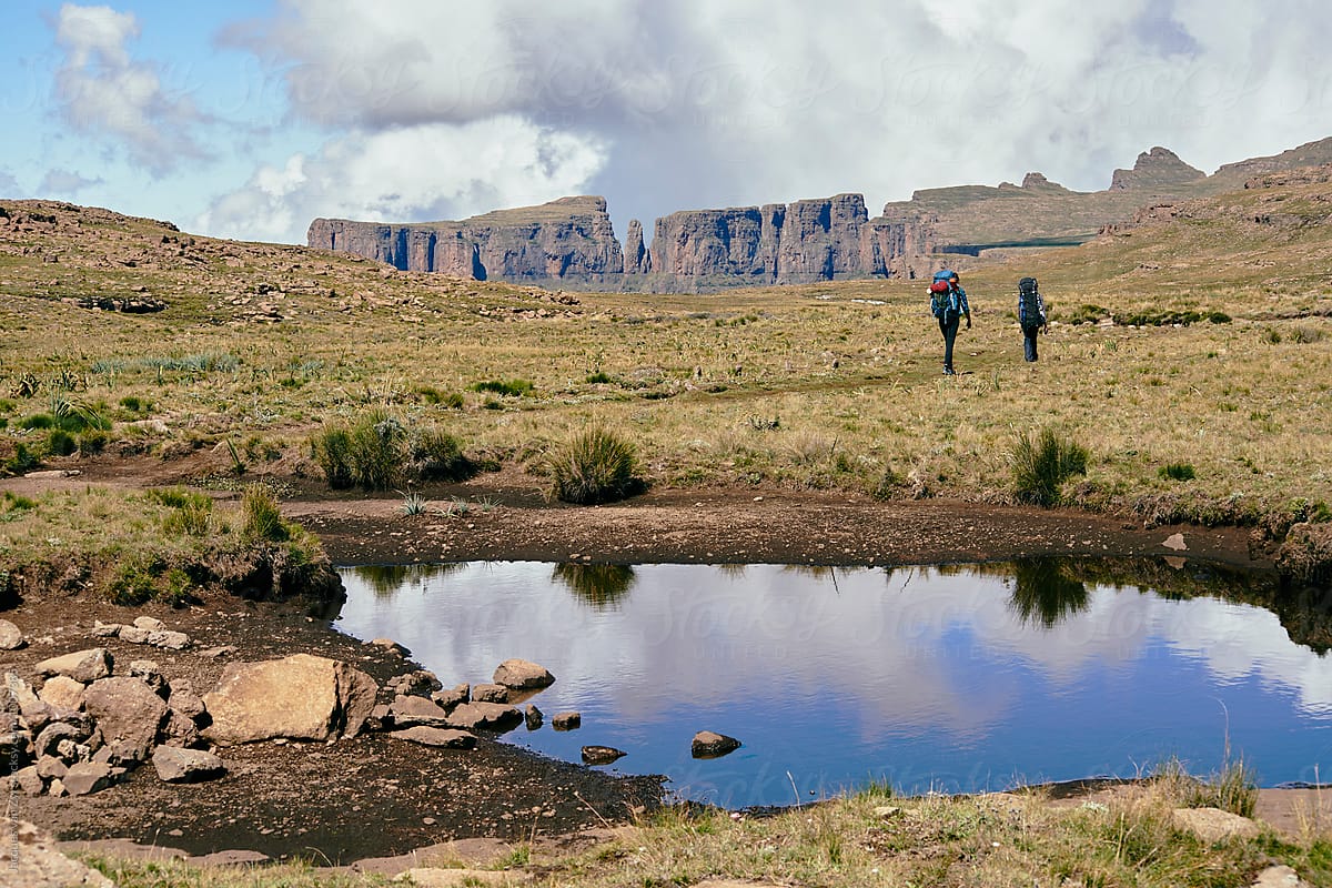 A pond reflecting cloud and sky with two hikers backpacking through mountains.
