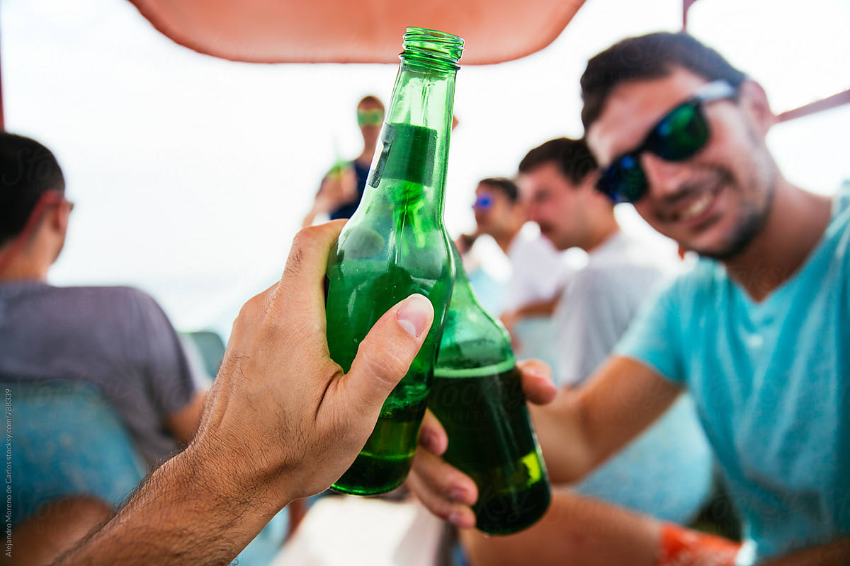 Front view of a man\'s hand holding a green beer bottle making a toast with a friend