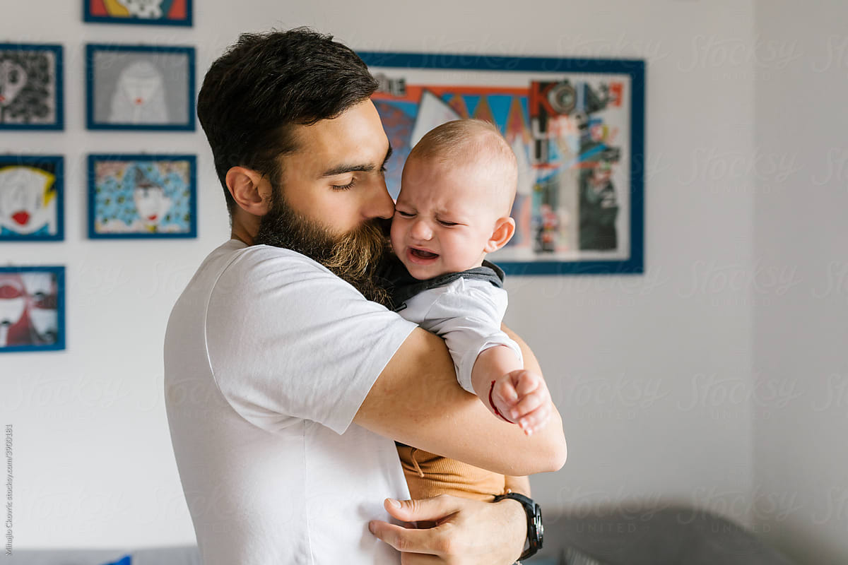 Crying baby being held by young bearded father at home