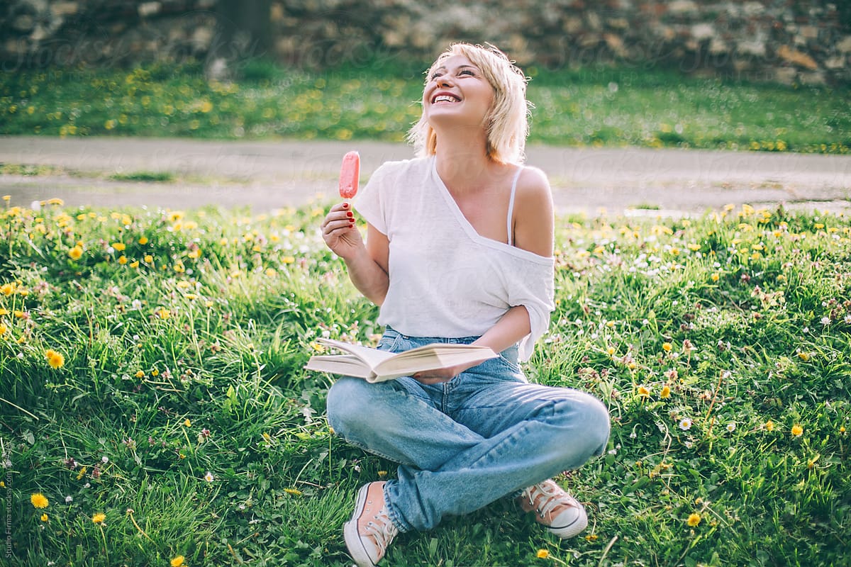 Woman sitting on the ground with ice-cream and book.