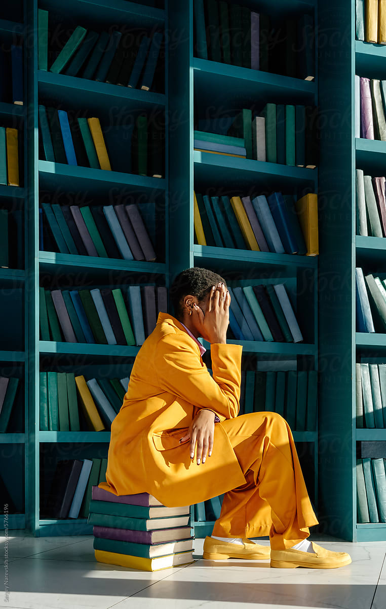 Anonymous person in suit sitting on stack of books near book shelves