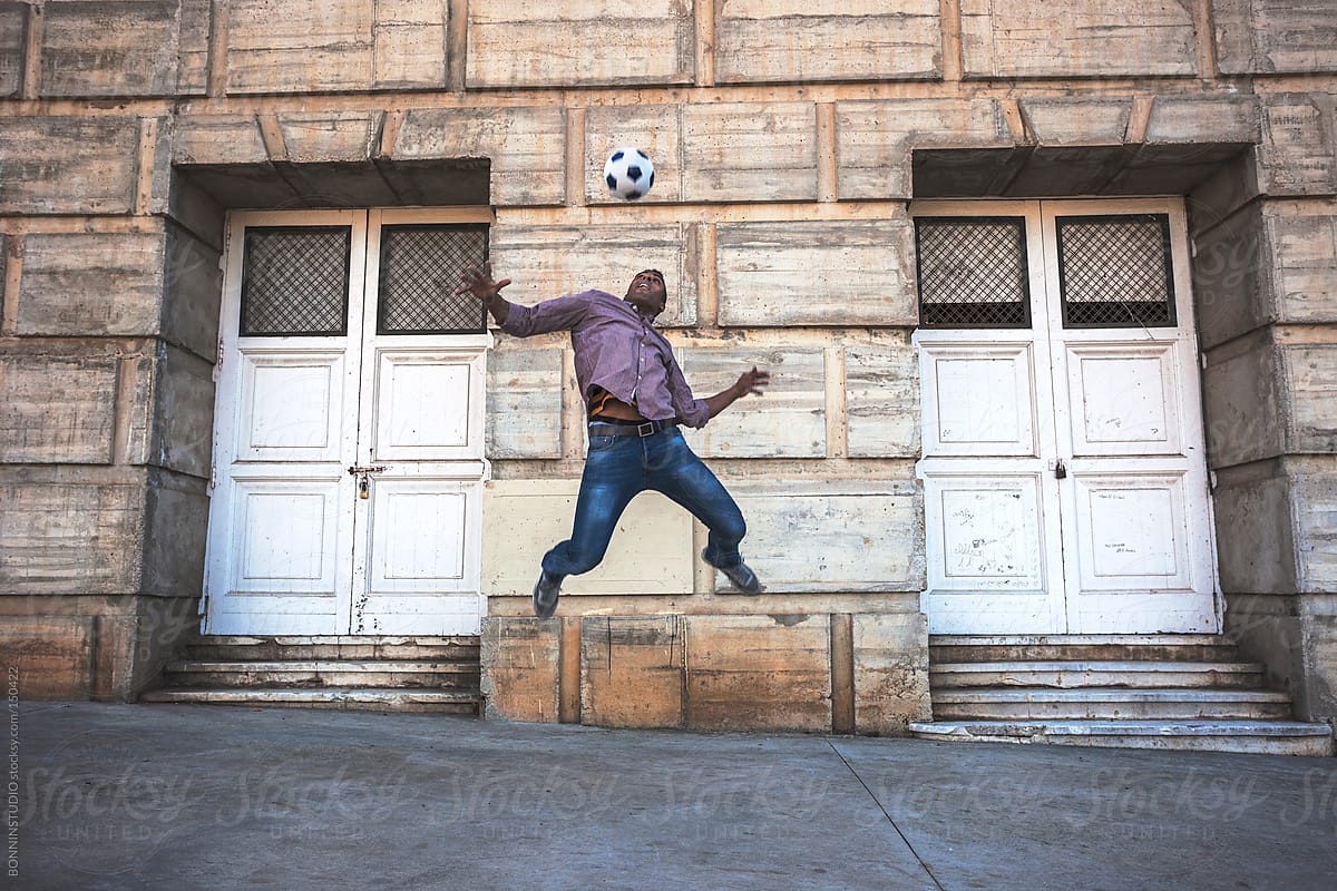 Black man jumping with soccer ball on the street in front two big white doors.