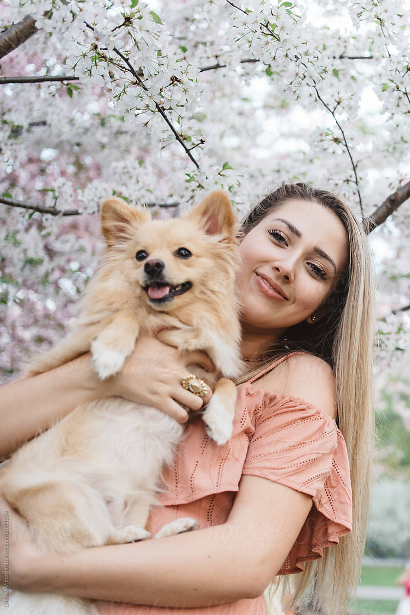 A young blonde woman standing amongst the cherry blossom trees with her dog